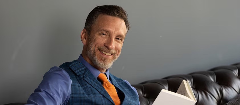 Mike Michalowicz, author of Profit First, smiling with the book open on a leather sofa